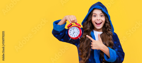 Happy child in hooded bathrobe pointing finger at alarm clock yellow background, time. Teenager child with clock alarm, horizontal poster. Banner header, copy space.