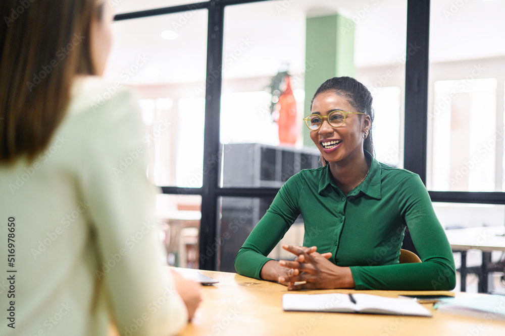Obraz premium Female manager praises young ambitious african-american female employee sitting in contemporary office space, proud young woman in glasses feels happy