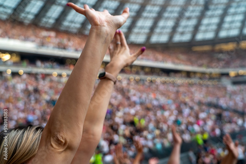 Raised hands of millennial woman with blurred crowd of people enjoying pop music festival in background. Many different people at concert listening to rock band music open air.
