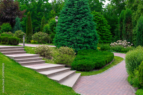 stone steps to path from paving slabs in park on slope with ornamental plants, evergreen bushes and trees, backyard landscaping with green lawn place for walk, iron lantern and drain system, nobody.