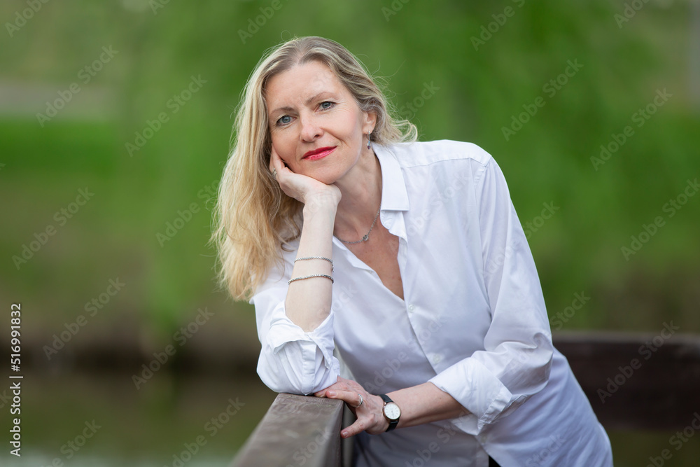 A beautiful middle-aged European woman. Fifty-year-old blonde woman in a white shirt on the background of nature. A beautiful middle-aged European woman. Fifty-year-old blonde woman in a white shirt on the background of nature.