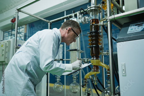 Caucasian biochemical scientist with lab coat, gloves and glasses is holding paperwork and recording the pressure and temperature of cannabis extraction. Marijuana researcher looking at this machine.