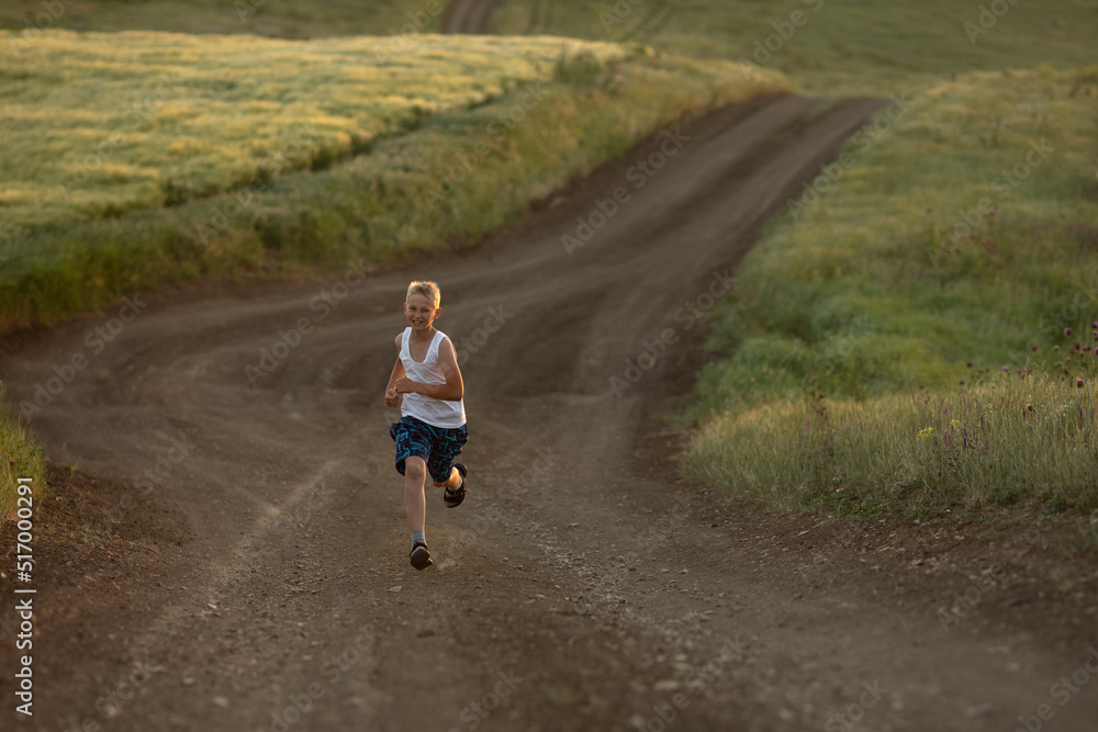 Obraz premium A boy in white t-shirt runs at sunset along a rural road next to a wheat field. Summer holidays in the countryside. Happy childhood