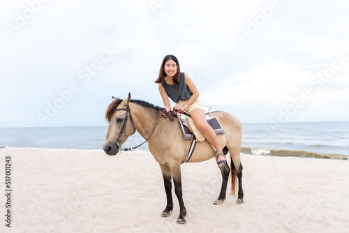 A young Asian woman is walking happily on a horse on the sandy beach before the sun goes down. Tourists come to relax at the beach, ride horses, and take a walk.