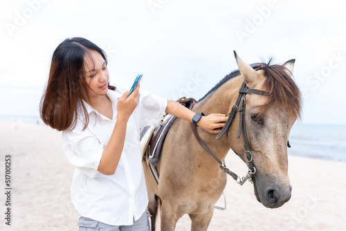 A young Asian woman in a white shirt with long hair is playing with the water on the beach in a tourist area on vacation. woman using mobile phone to take pictures of happy horses © Ekkasit A Siam