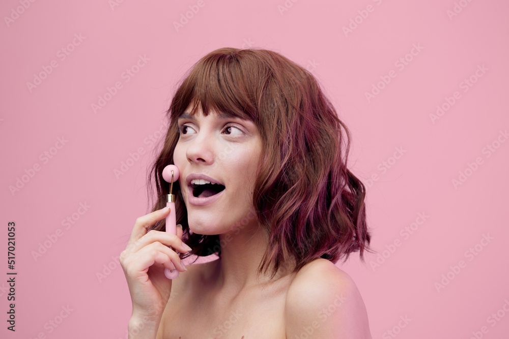 a happy, enthusiastic woman, standing on a pink background with pink lighting from the side, with her hair styled, doing a facial massage with a pink roller, her mouth wide open in surprise. a happy, enthusiastic woman, standing on a pink background with pink lighting from the side, with her hair styled, doing a facial massage with a pink roller, her mouth wide open in surprise.