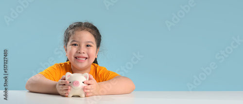 Asian cute girl saving money putting coins into piggy bank on table at home, learning about saving, Kid save money for future education. Money, finances, insurance, and people concept