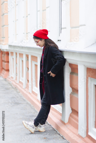 A young woman wearing a red hat, red scarf and a black coat is leaning on the white and orange classic building at the street