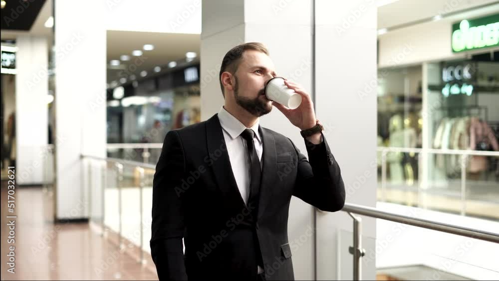 A young man with a beard dressed in a business suit and tie is drinking coffee in a shopping and business center. Business man with a glass of coffee. A young man with a beard dressed in a business suit and tie is drinking coffee in a shopping and business center. Business man with a glass of coffee.
