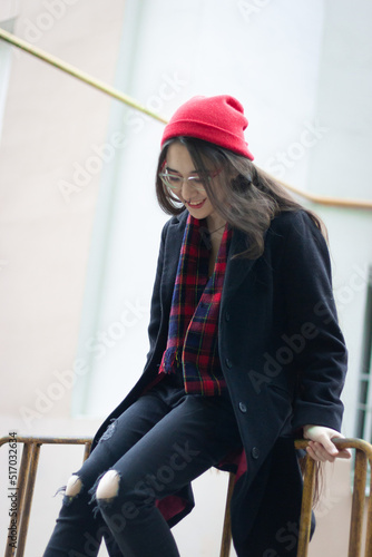 A young woman wearing a red hat, red scarf and a black coat is sitting on a green fance against the white and orange classic building at the street