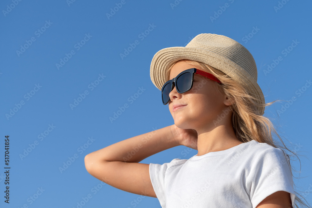 A young girl in a hat and sunglasses straightens her hair with one hand against the blue sky.