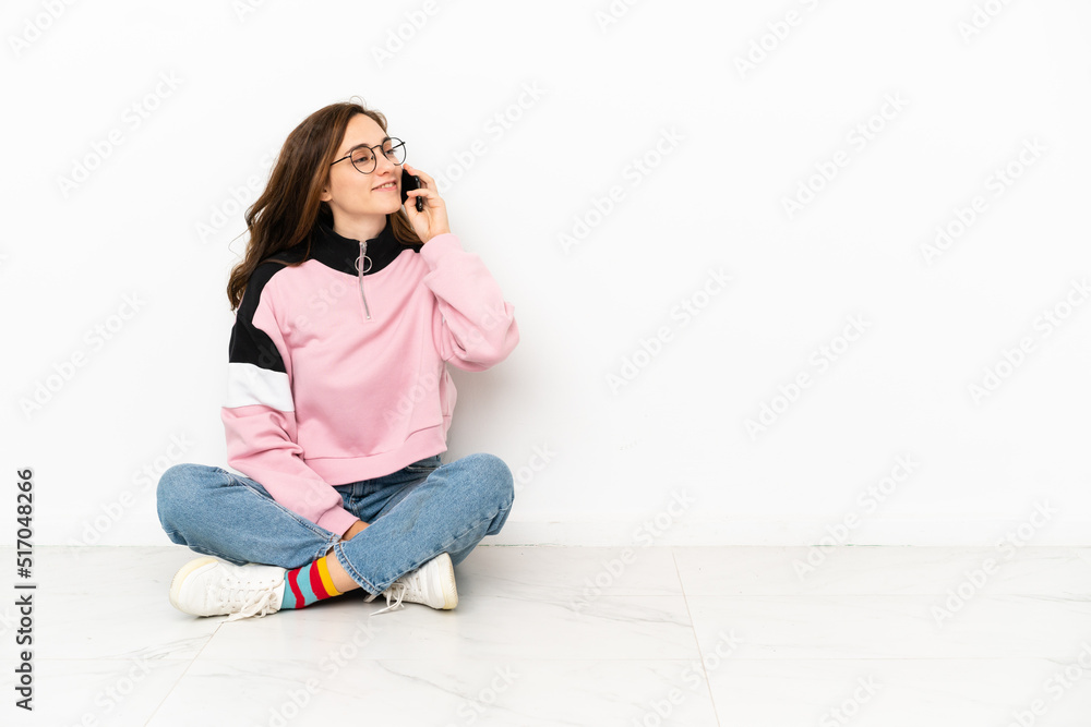 Young caucasian woman sitting on the floor isolated on white background keeping a conversation with the mobile phone