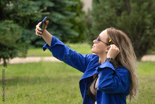 a beautiful blonde with long wavy hair in a blue shirt on a walk in the park, in her hands is a phone on which she takes a selfie. Young girl on vacation taking a picture of herself
