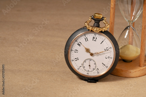 Antique pocket watch and glass hourglass on beige background with space for text, place for presentation. Round grey retro clock with golden hands and white dial.
