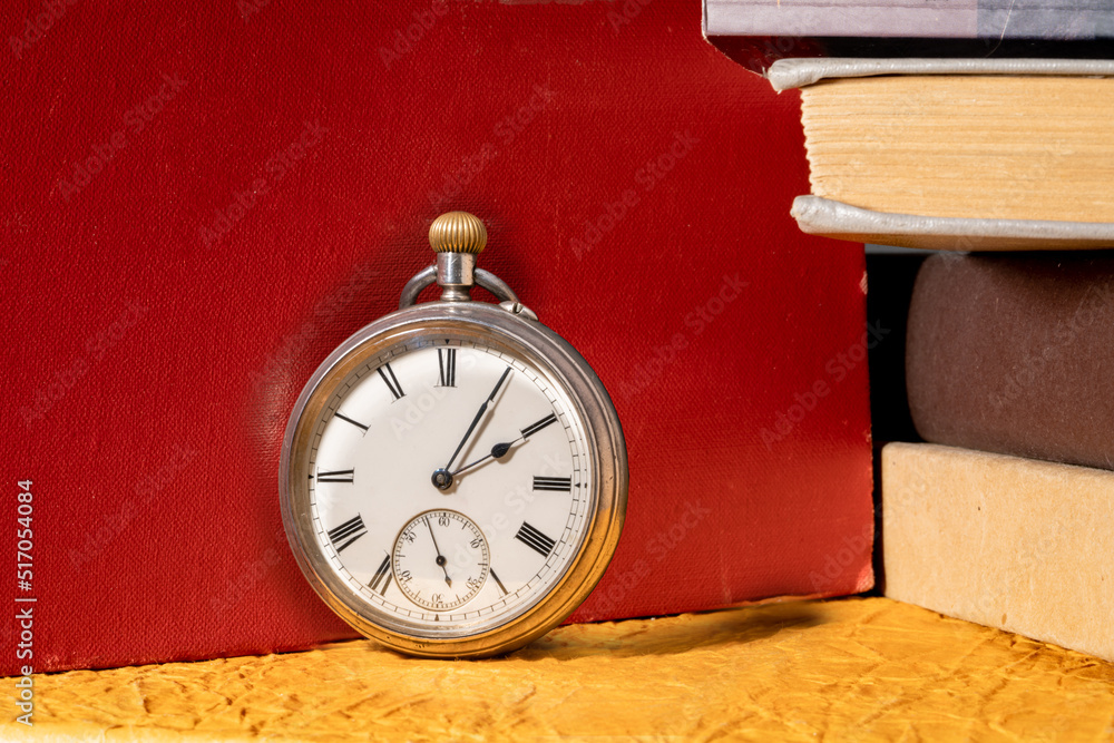 Antique silver retro clock on background of stacks of books in library. Round old vintage clock with dial and hands near books with red, yellow and gray covers. Antique silver retro clock on background of stacks of books in library. Round old vintage clock with dial and hands near books with red, yellow and gray covers.