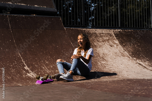 Happy smiling girl with skate board sitting on skate playground and having fun. Extreme sport lifestyle. Laughing child with skate board posing on sport ramp.