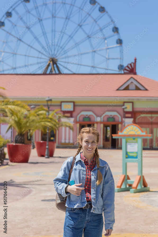 A cheerful woman with pigtails in a denim outfit walks in a sunny amusement park against the backdrop of a ferris wheel. Weekend for a walk