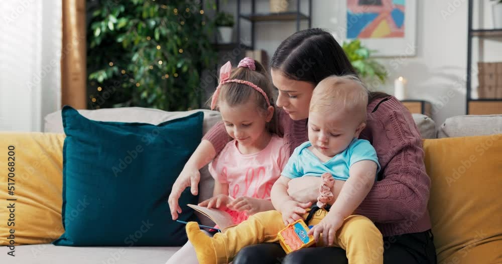 Happy family young mother babysitter hold read book relax embrace cute little children daughters, smiling parent mum tell small kids funny fairy tale story sit on sofa having fun together at home. Happy family young mother babysitter hold read book relax embrace cute little children daughters, smiling parent mum tell small kids funny fairy tale story sit on sofa having fun together at home.