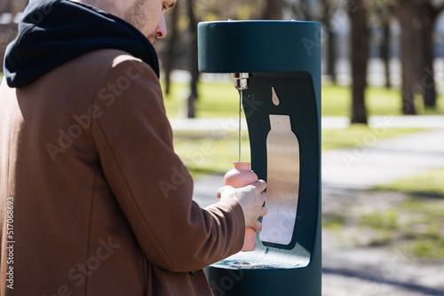Man refilling his water bottle at the city. Free public water bottle refill station. Sustainable and green city. Male in brown coat.  Tap water to reduce plastic bottle usage. Drinking water dispenser photo