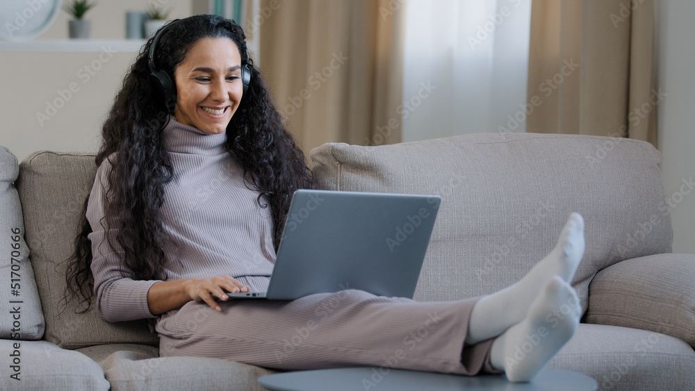 Hispanic arabian cute woman girl wear headphones sit on sofa at home living room hold laptop use video call talk distance wave hand greet boyfriend smiling social network communication during pandemic Hispanic arabian cute woman girl wear headphones sit on sofa at home living room hold laptop use video call talk distance wave hand greet boyfriend smiling social network communication during pandemic