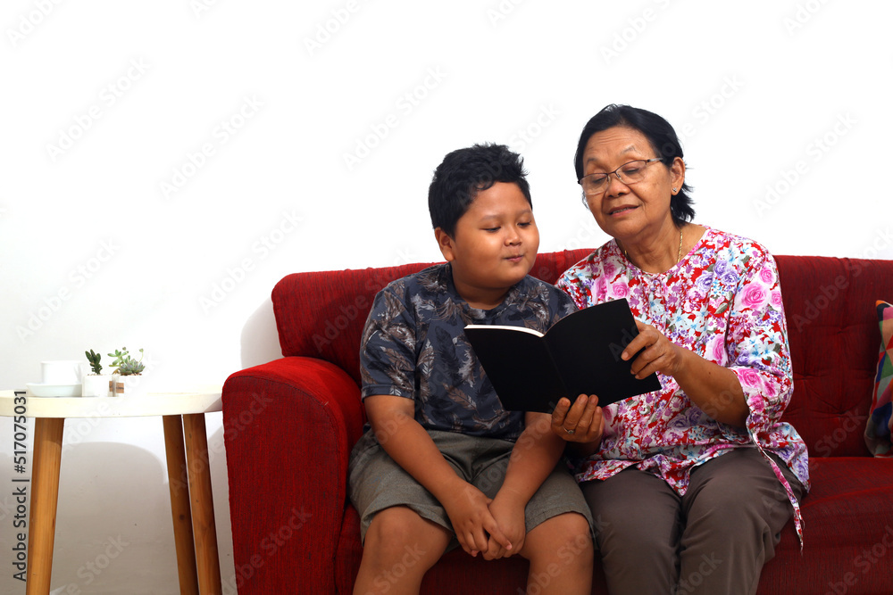 Young asian boy and asian grandmother reading book at home. Front view with copyspace