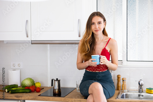 Portrait of attractive woman sitting on kitchen table and drinking coffee.