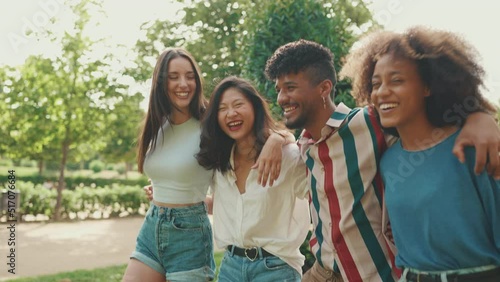 Happy multiethnic young people walk embracing on summer day outdoors. Group of friends are talking and laughing merrily while walking along path in city park