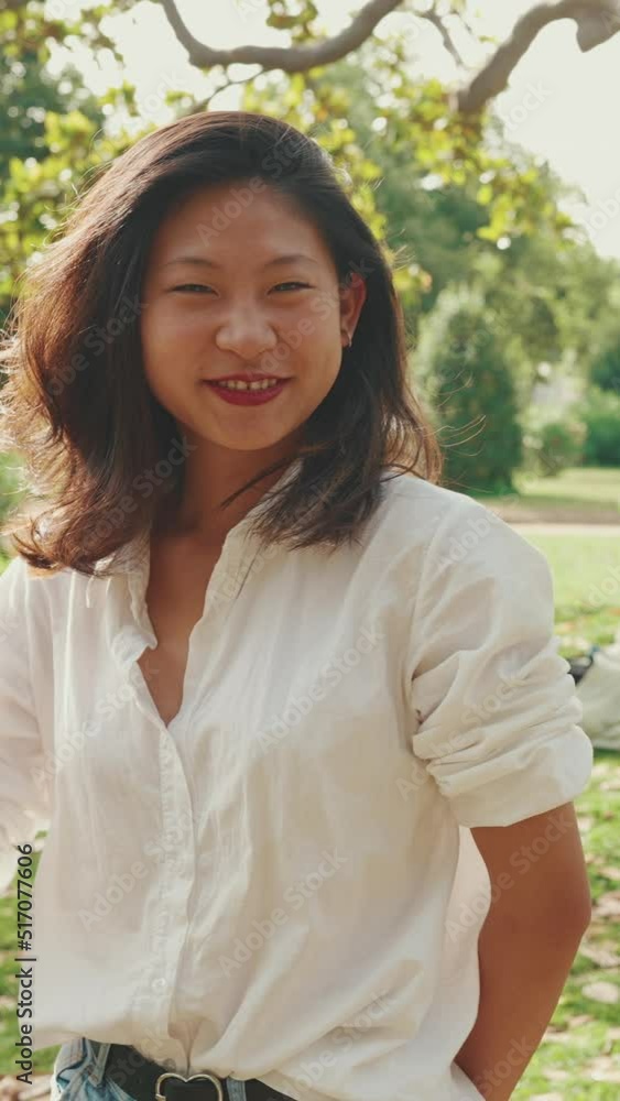 VERTICAL VIDEO: Young Asian smiling woman with long brown hair wearing white t-shirt posing for the camera in the park . Picnic on summer day outdoors VERTICAL VIDEO: Young Asian smiling woman with long brown hair wearing white t-shirt posing for the camera in the park . Picnic on summer day outdoors