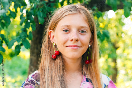 Defocus beautiful blond teen girl with sweet cherry on ear. Beautiful smiling teenage girl closeup, against green of summer park. Child earrings. Summer portrait. Out of focus photo