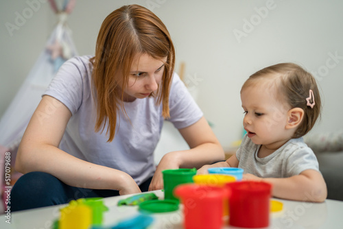 One girl small caucasian toddler child playing with colorful plasticine on the table at home with her mother woman childhood and growing up development concept copy space side view