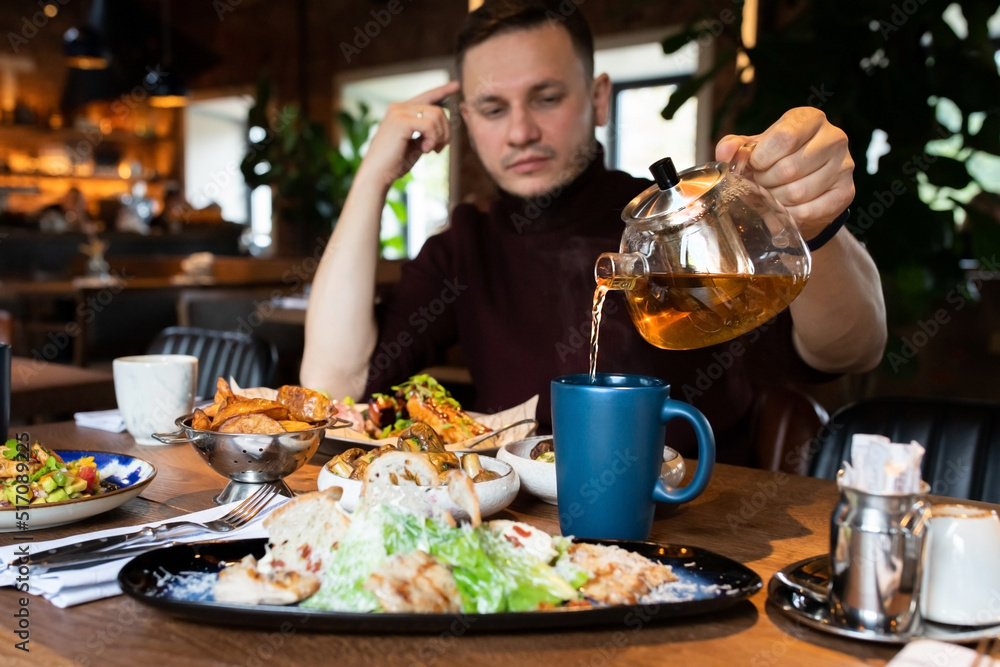A man in a restaurant pours himself a mug of hot tea from a glass teapot. Front view.