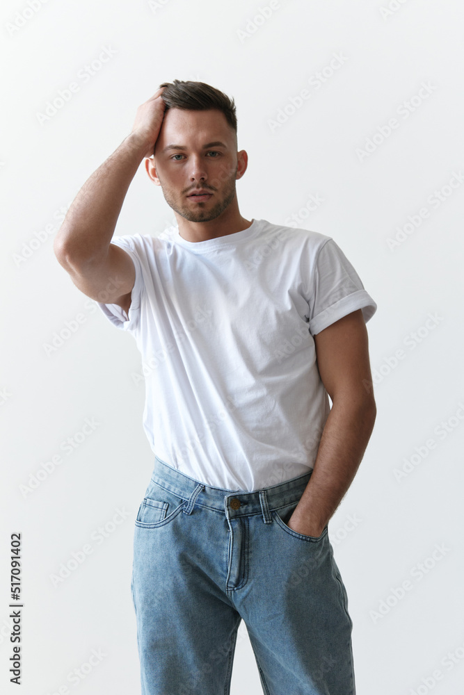 Fototapeta premium Portrait shot of handsome serious tanned man guy in basic t-shirt holds hand on head looks at camera posing on white background. Fashion Style New Collection Offer. Copy space for ad. Model snap
