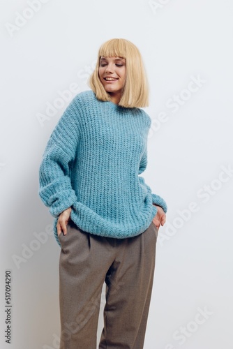 a stylish, happy blonde woman stands on a light background in a long blue sweater, smiling pleasantly and holding her hands in the pockets of brown pants