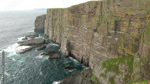 A slow tiliting shot reveals seabirds flying over a turquoise green ocean in front of a dramatic sea cliff rising straight up out of the ocean as waves crash against its base. Handa Island, Scotland. photo