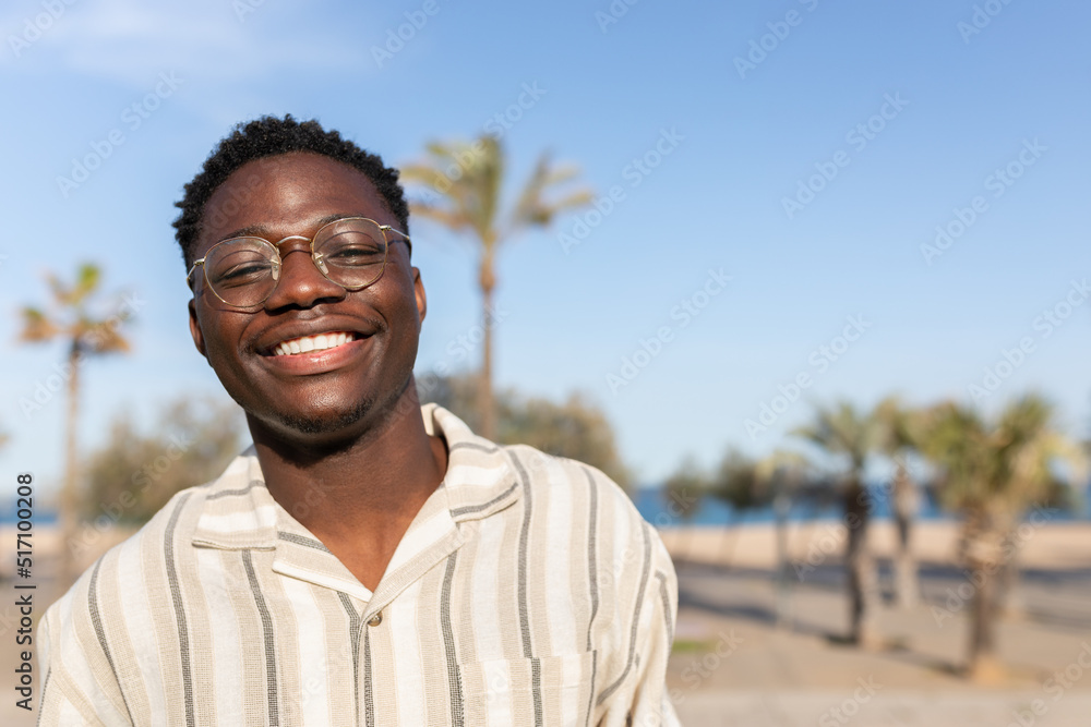Portrait of young happy and smiling black man wearing glasses outdoors. Background with palm trees. Copy space. Portrait of young happy and smiling black man wearing glasses outdoors. Background with palm trees. Copy space.