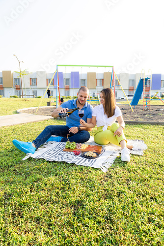 Beautiful Latin couple having a romantic breakfast with lots of tasty food and wine, sitting together on the picnic blanket in the park on a sunny morning.