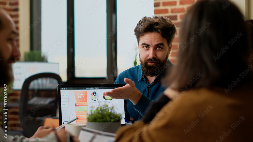 Project manager pointing at business charts on laptop, showing online research data to colleagues. People planning startup project and presentation with charts, creating successful report. Project manager pointing at business charts on laptop, showing online research data to colleagues. People planning startup project and presentation with charts, creating successful report.