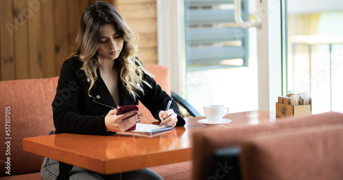 Serious female student concentrated on writing plan in notepad while sending messages on phone sitting at cafe, pensive girl noting information from smartphone