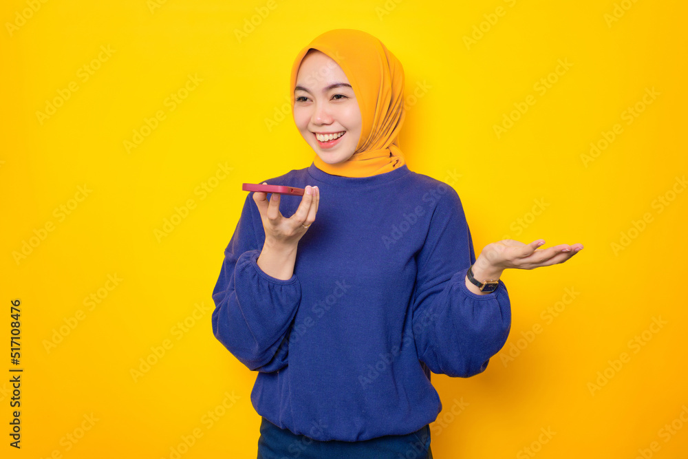 Cheerful young Asian Muslim woman dressed in casual sweater talking on smartphone using loudspeaker mode isolated over yellow background Cheerful young Asian Muslim woman dressed in casual sweater talking on smartphone using loudspeaker mode isolated over yellow background