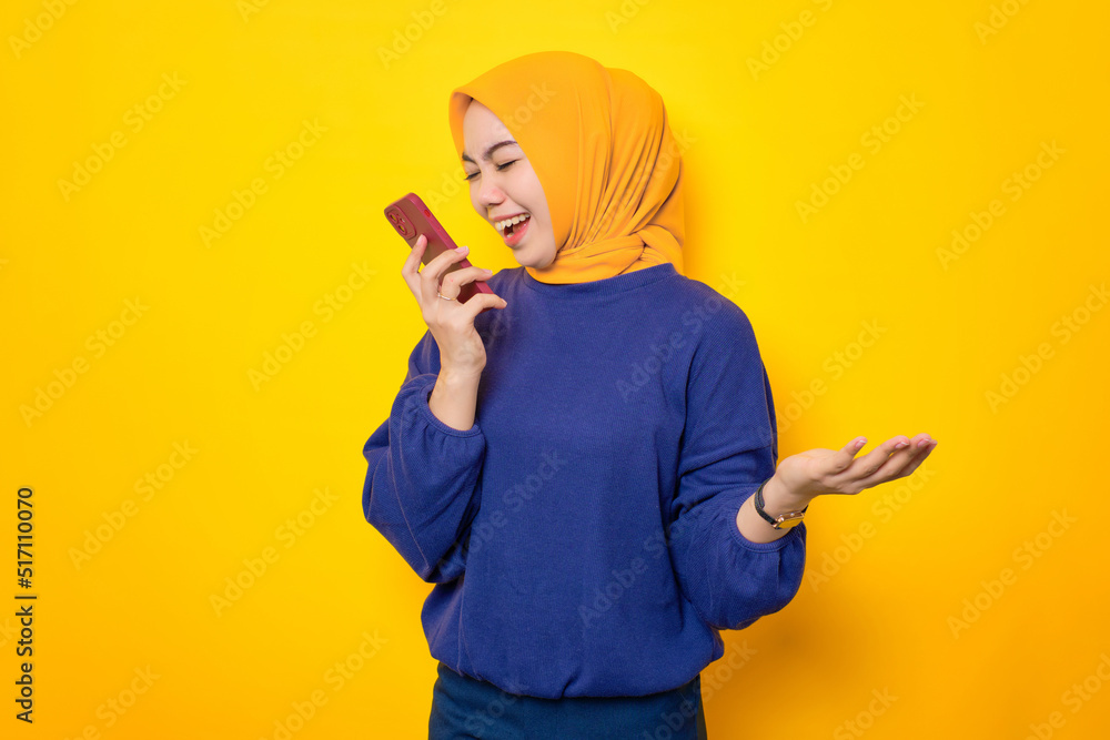 Angry young Asian Muslim woman dressed in casual sweater shouting at mobile phone, reacting to bad news isolated over yellow background Angry young Asian Muslim woman dressed in casual sweater shouting at mobile phone, reacting to bad news isolated over yellow background