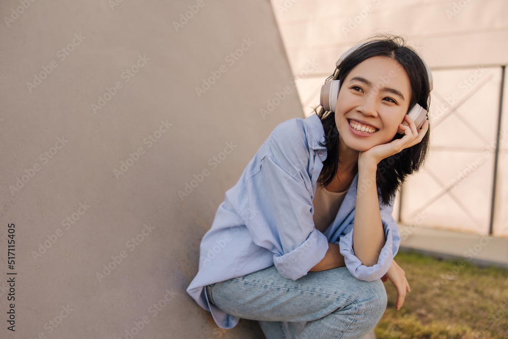 Cheerful asian pretty woman looking away with smile listening music. Brunette in headphones sitting near to wall in the city. Lifestyle, relaxing concept Cheerful asian pretty woman looking away with smile listening music. Brunette in headphones sitting near to wall in the city. Lifestyle, relaxing concept