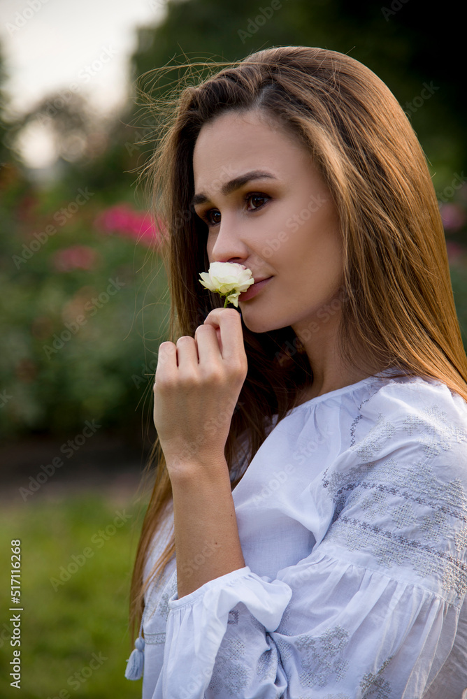 portrait of a beautiful brunette smiling woman in white embroidered shirt smelling a fragrant white rose flower in a summer garden portrait of a beautiful brunette smiling woman in white embroidered shirt smelling a fragrant white rose flower in a summer garden