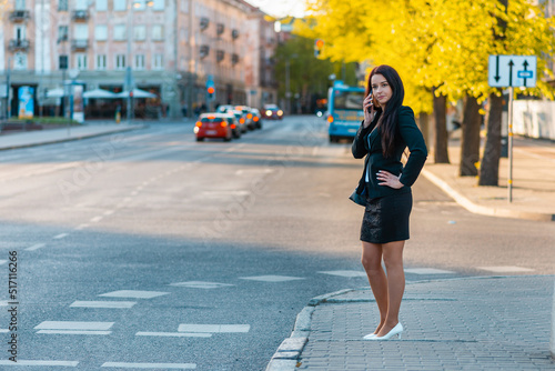 Young business woman with smartphone and standing against street blurred building background. Fashion business photo of beautiful girl in black suite with phone. Closeup.