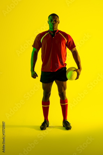 Portrait of african american male rugby player with rugby ball over yellow lighting