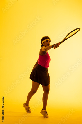 Vertical image of african american female tennis player hitting ball in yellow lighting