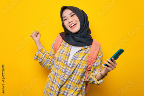 Excited young Asian Muslim woman student in Plaid shirt and backpack using mobile phone and doing winner gesture isolated on yellow background. Education school university college concept