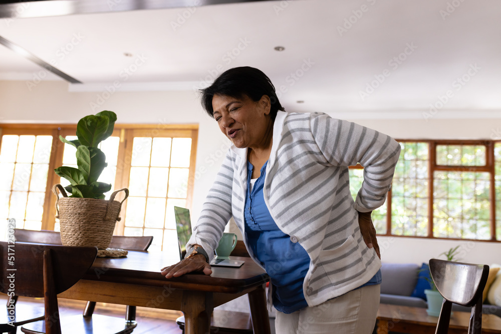 Biracial mature woman with backache touching back and standing by wooden table at home Biracial mature woman with backache touching back and standing by wooden table at home