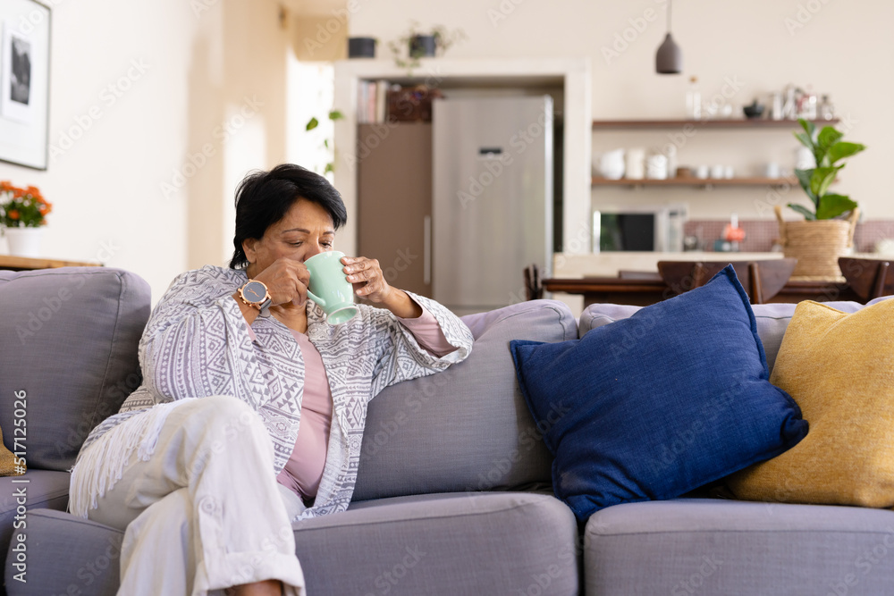 Biracial mature woman with short hair drinking coffee while relaxing on sofa in living room Biracial mature woman with short hair drinking coffee while relaxing on sofa in living room