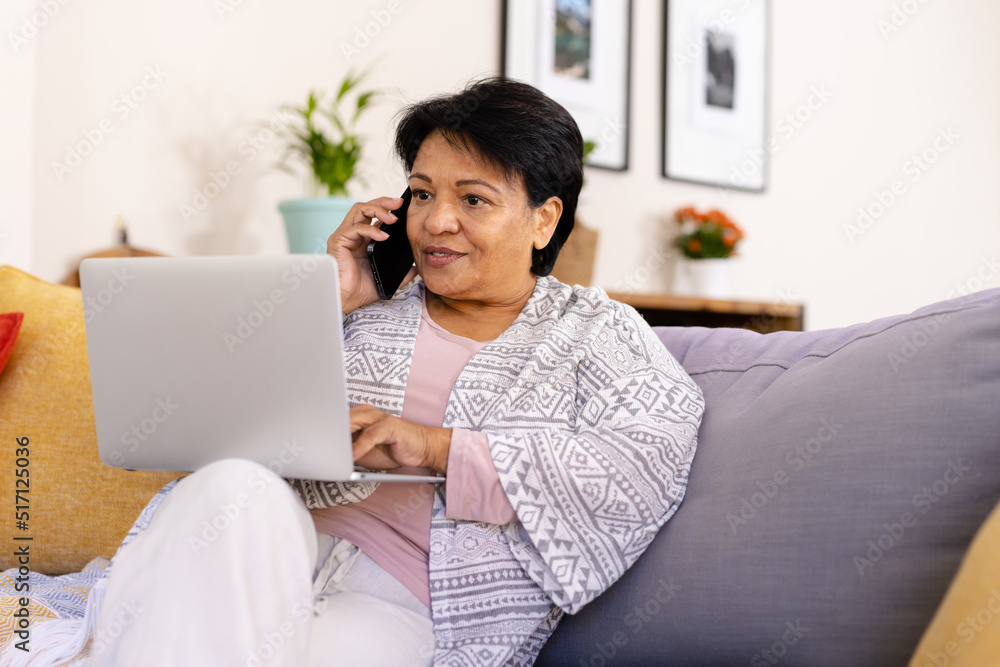 Biracial mature woman with short hair talking over cellphone and using laptop while relaxing on sofa Biracial mature woman with short hair talking over cellphone and using laptop while relaxing on sofa