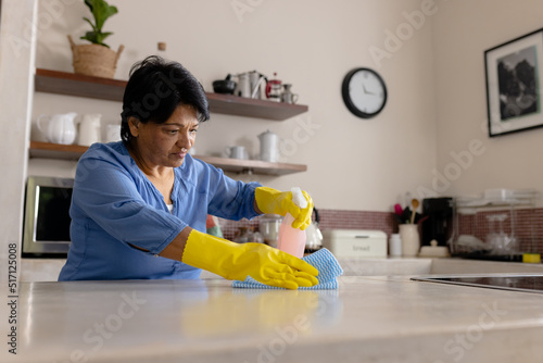 Biracial mature woman wearing gloves cleaning kitchen island with rag and spray bottle, copy space photo